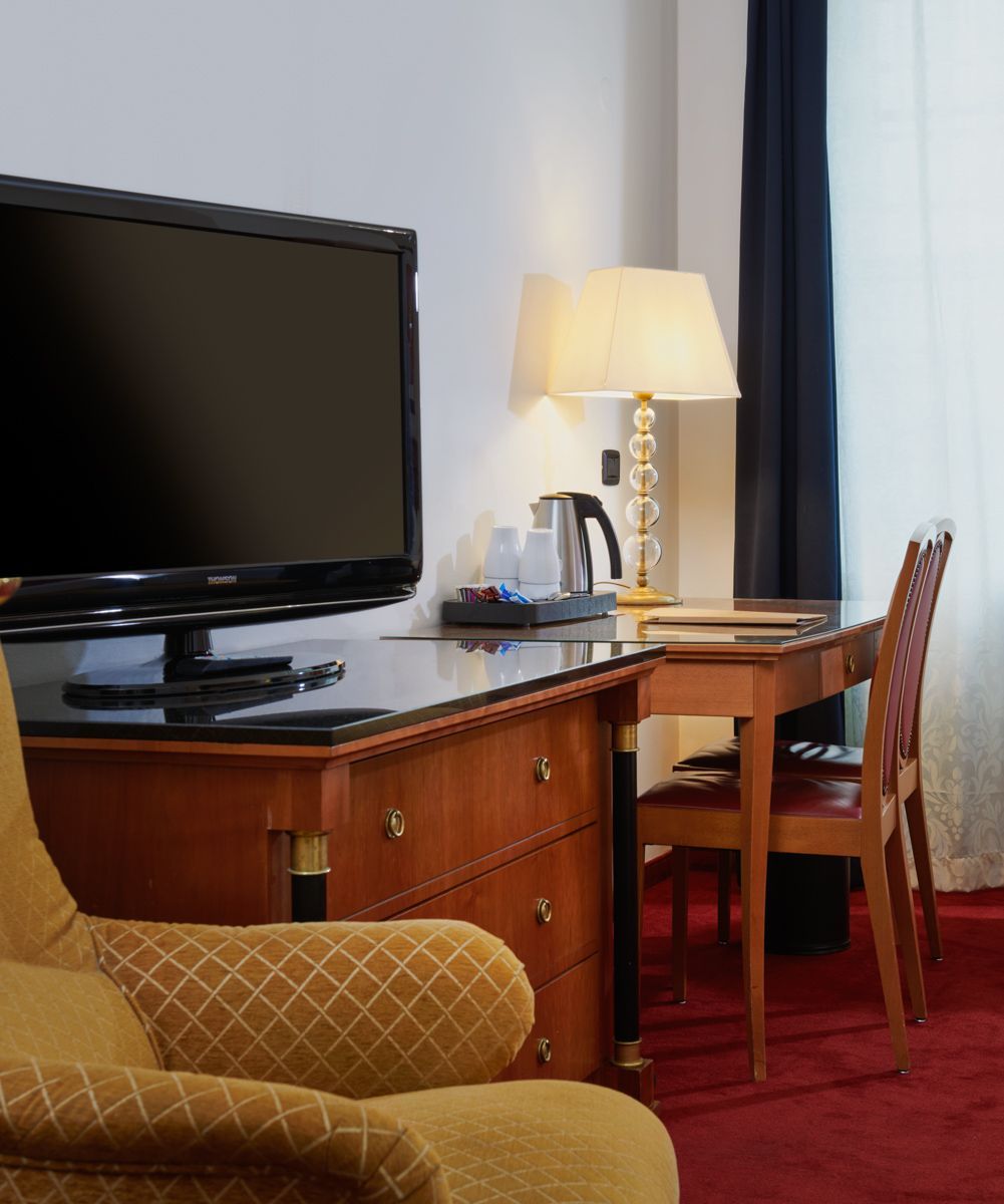 Camera d'albergo: TV su una cassettiera, scrivania con sedia, lampada. Tappeto rosso e tenda sullo sfondo.