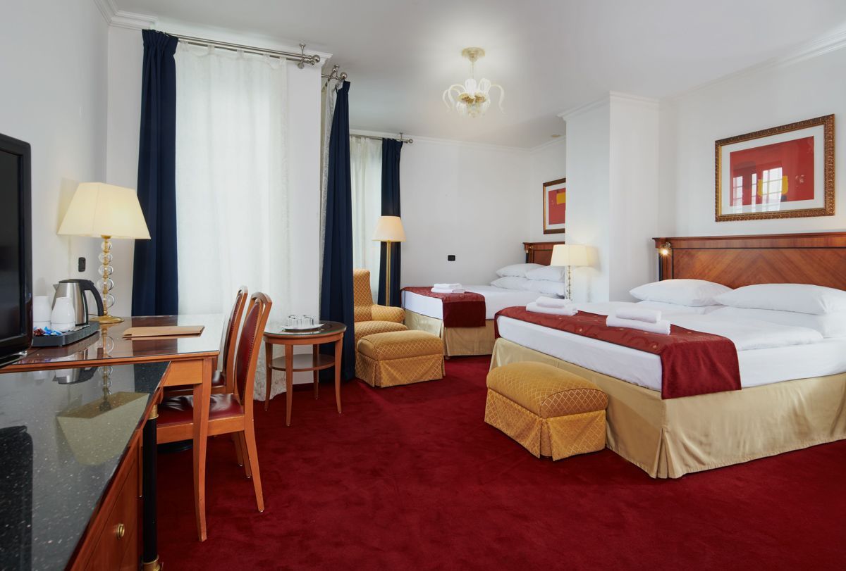 Camera d'albergo con due letti, tappeto rosso, scrivania, sedia, finestra e piccola area salotto.