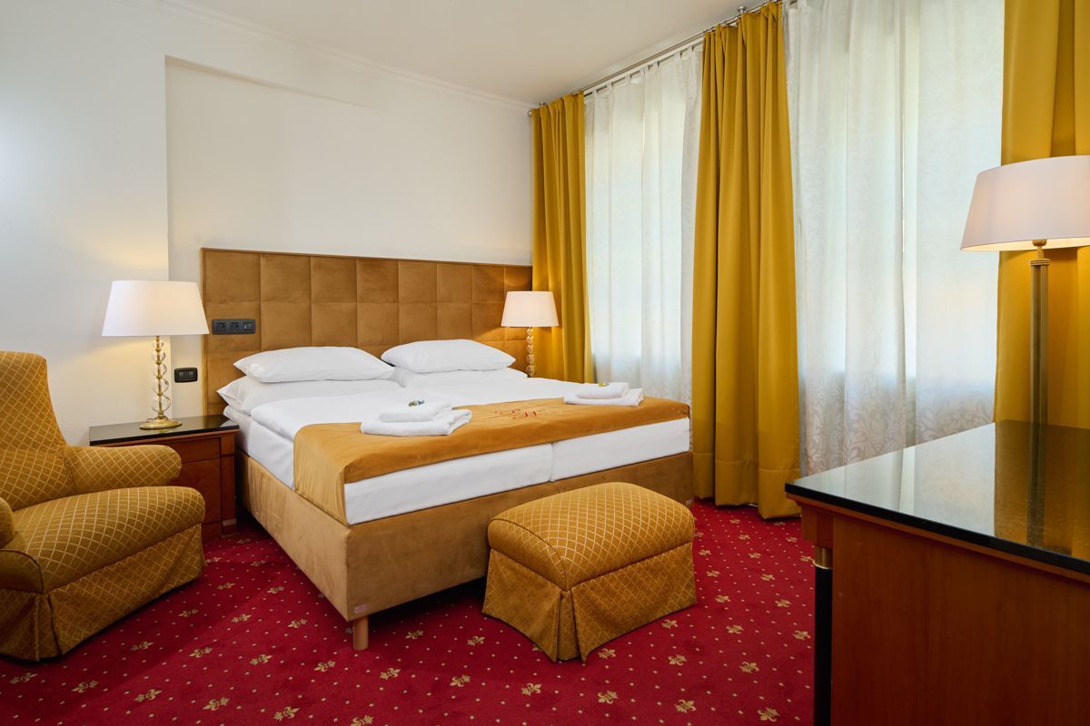 Camera d'albergo con letto, sedia, pouf, lampade, tende gialle e tappeto rosso.