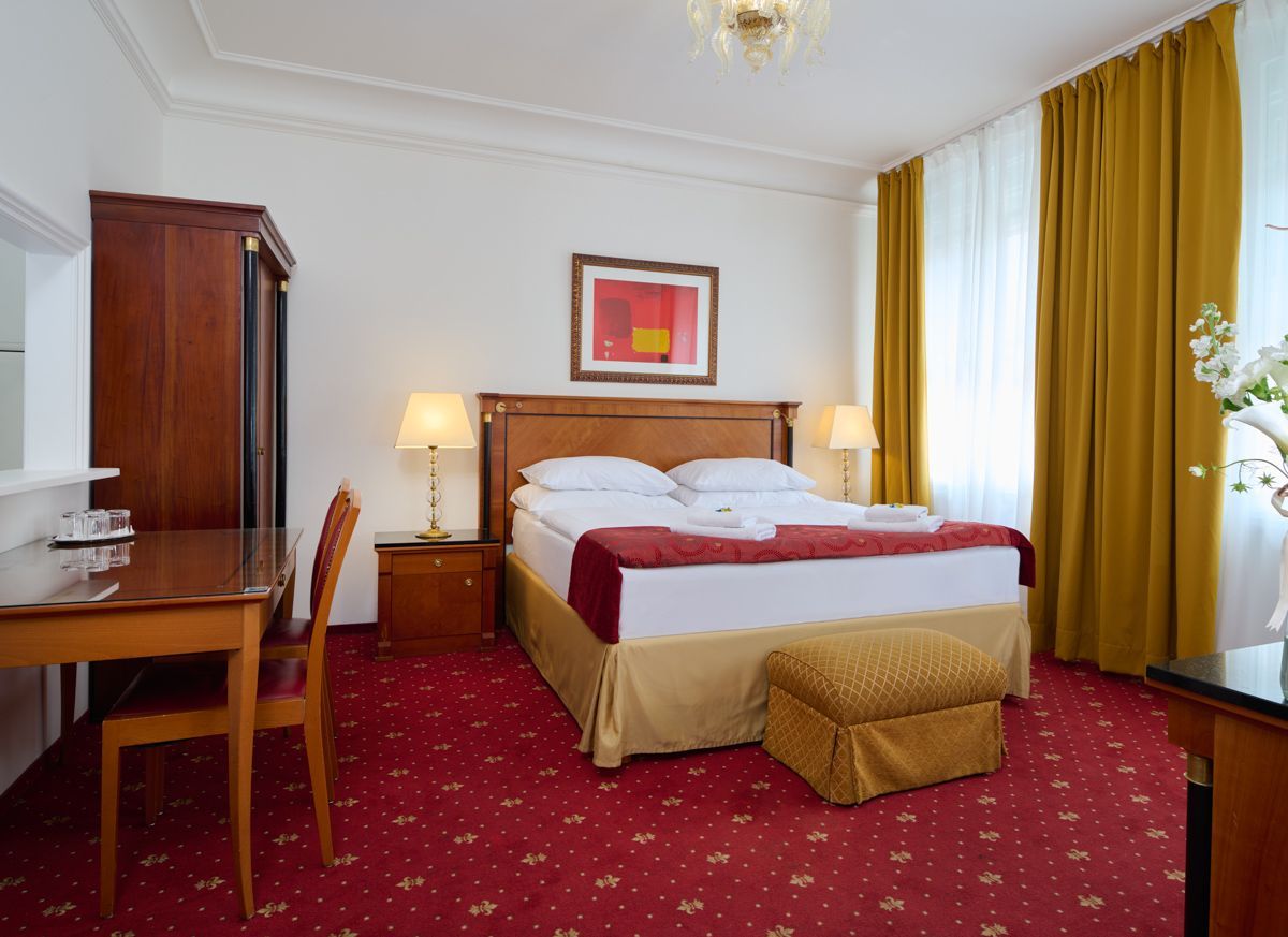 Camera d'albergo con letto, scrivania e armadio su un tappeto rosso a motivi geometrici; tende dorate e pareti color crema.