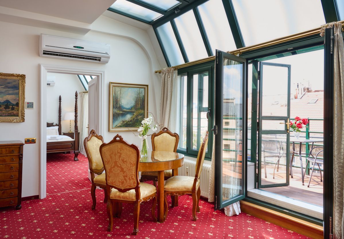 Stanza con tappeto rosso, tavolo e sedie, porte aperte su un balcone e uno scorcio di una camera da letto.