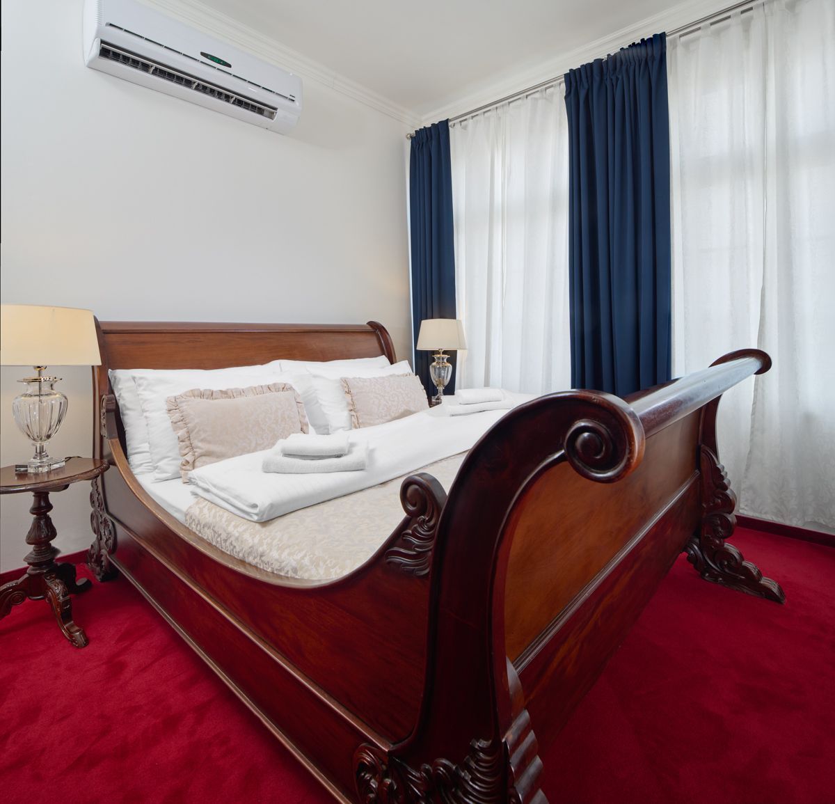 Camera da letto con letto in legno decorato, tappeto rosso, pareti bianche, tende blu e due lampade.