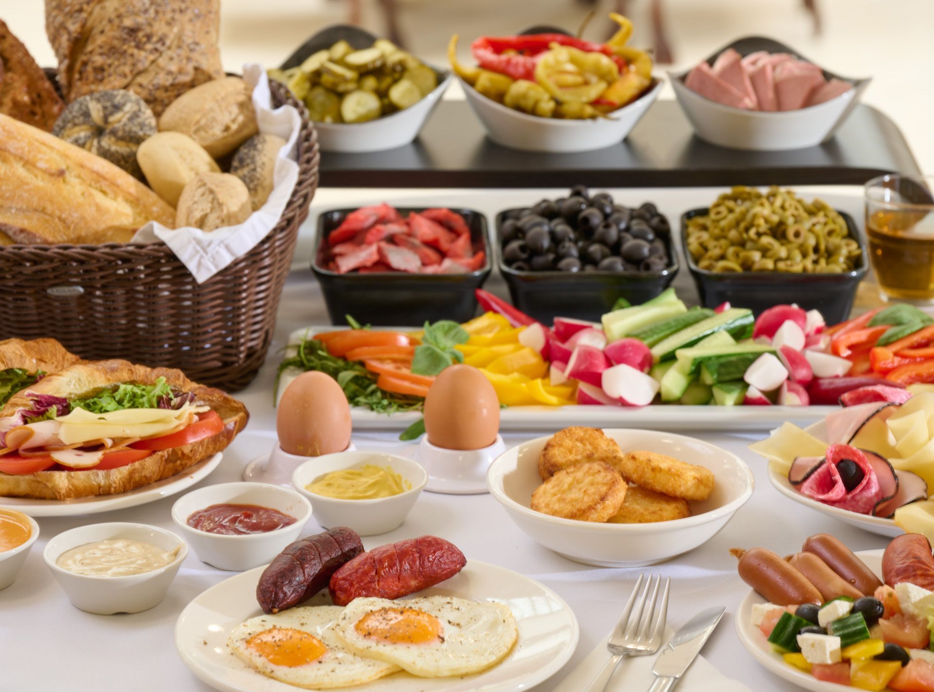 Colazione a buffet: pane, uova, salsicce, insalate e verdure sottaceto servite su una tovaglia bianca.