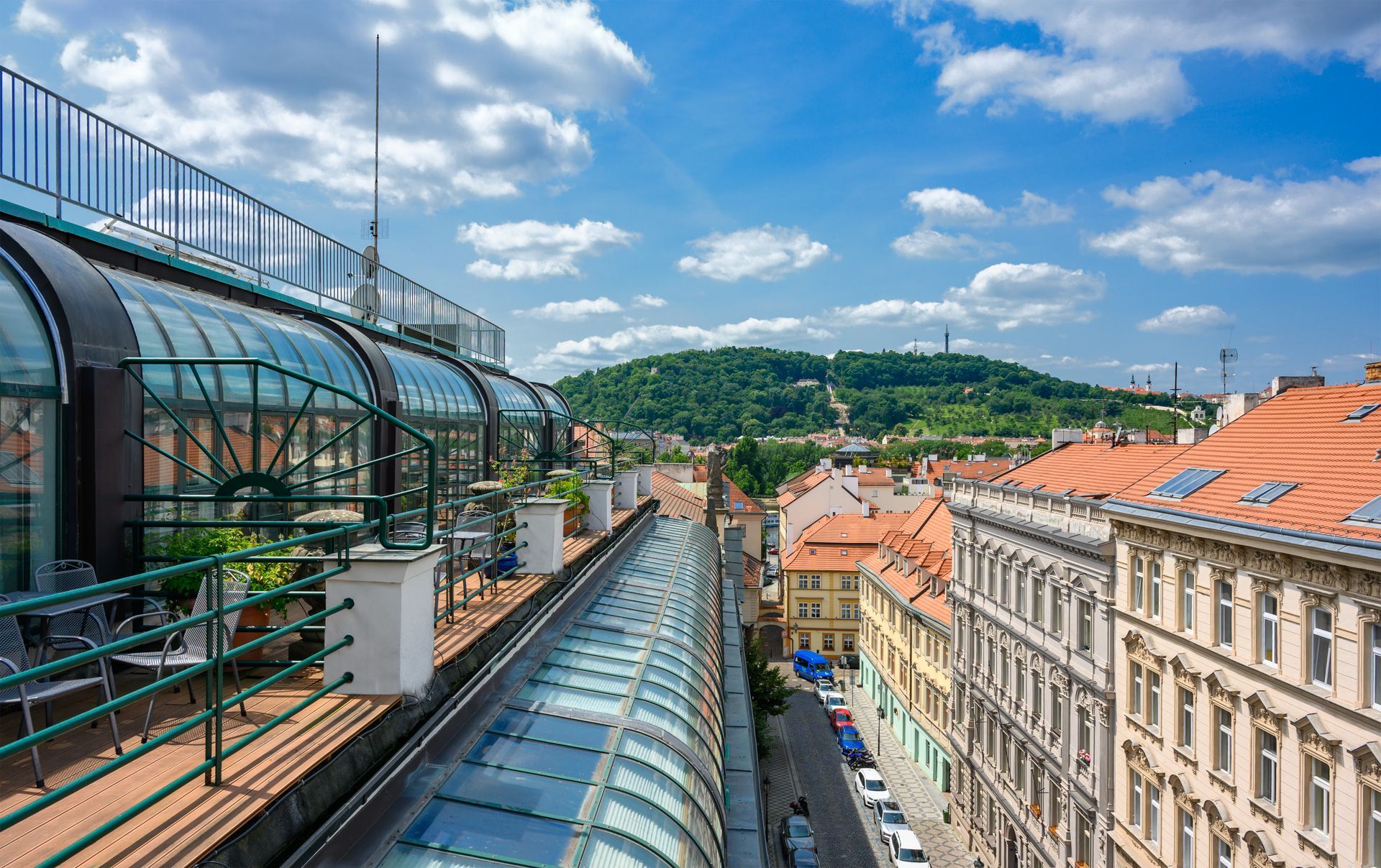 Vista panoramica di una città europea con una passerella coperta di vetro, edifici con tetti di tegole rosse e una collina sullo sfondo sotto un cielo azzurro.