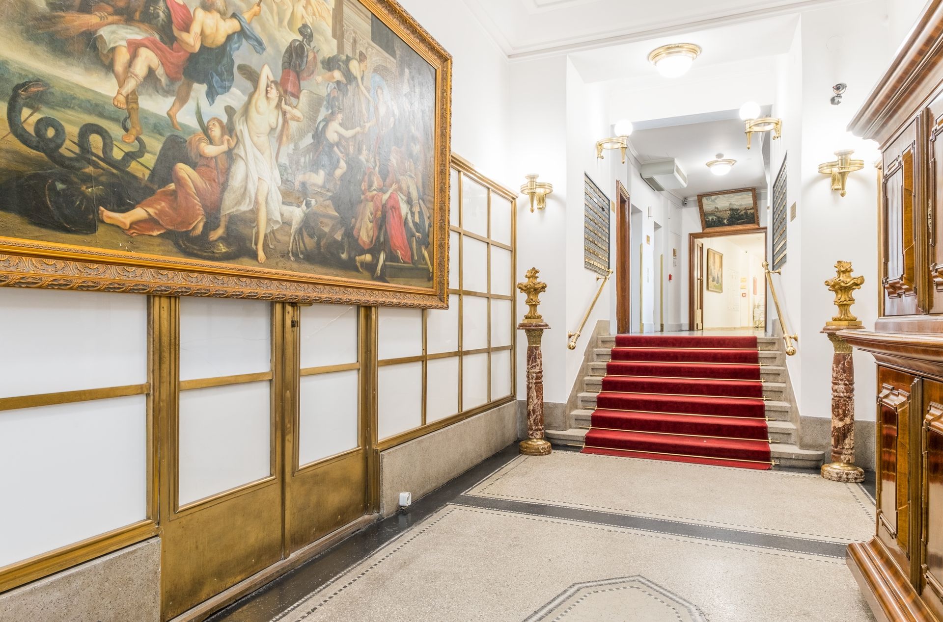 Ampio corridoio con grandi dipinti, scale con moquette rossa, dettagli dorati e pavimento decorato.