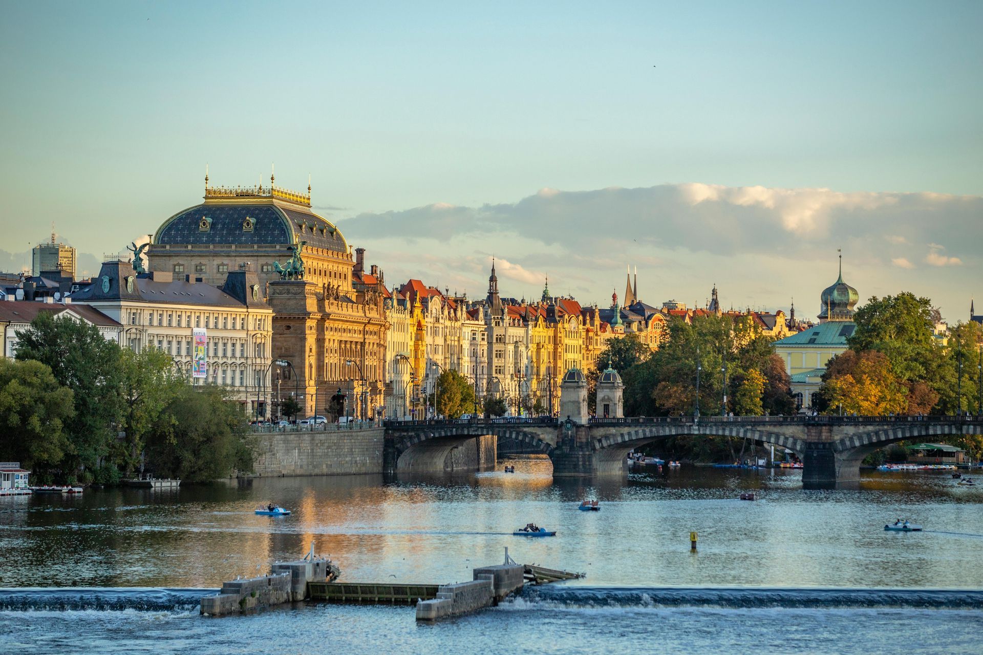 Prager Stadtbild bei Sonnenuntergang. Gebäude säumen den Fluss, darunter ein großes Theater mit Kuppeldach.