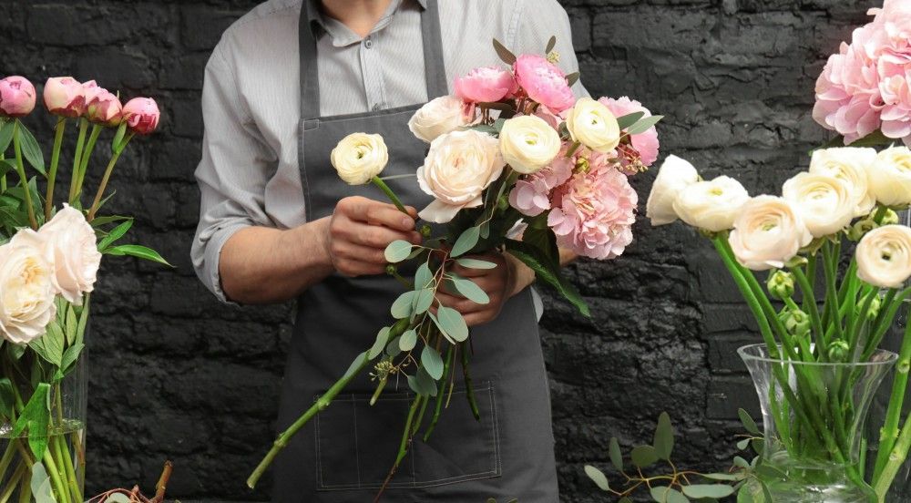 Fioraio che prepara un bouquet di fiori rosa chiaro e crema, grembiule scuro, parete di sfondo nera.