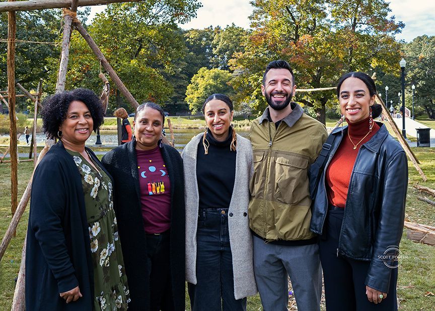 Five people pose outdoors; trees in background. Two women on either side, three in the middle.