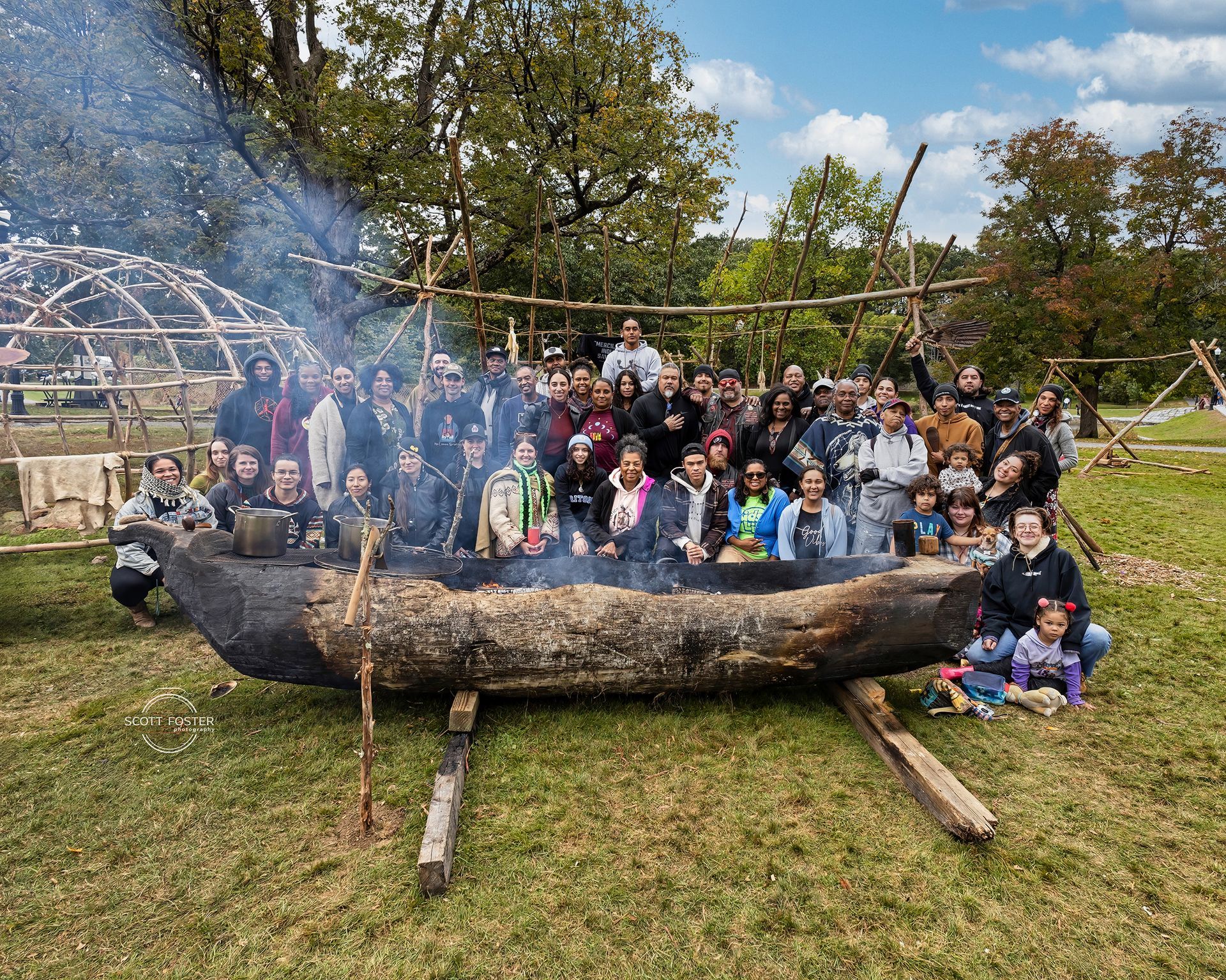 Group of people around a large, wooden canoe with smoke, near a wooden structure, in a park.