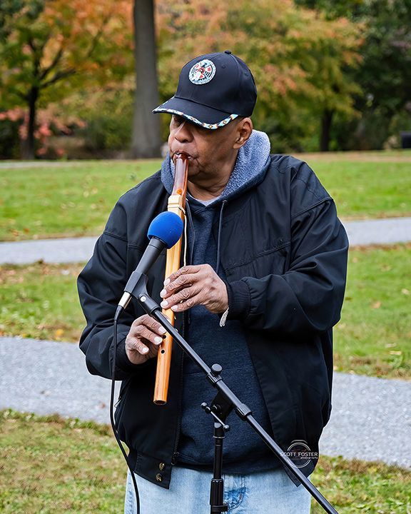 Man playing a wooden flute into a microphone outdoors; wearing a baseball cap and jacket.