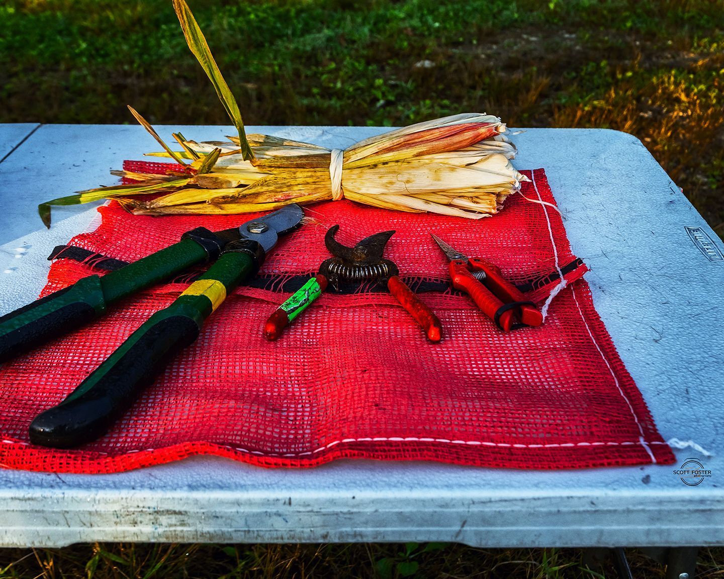 Gardening tools on red netting, with bound dried plants on a white table outdoors.