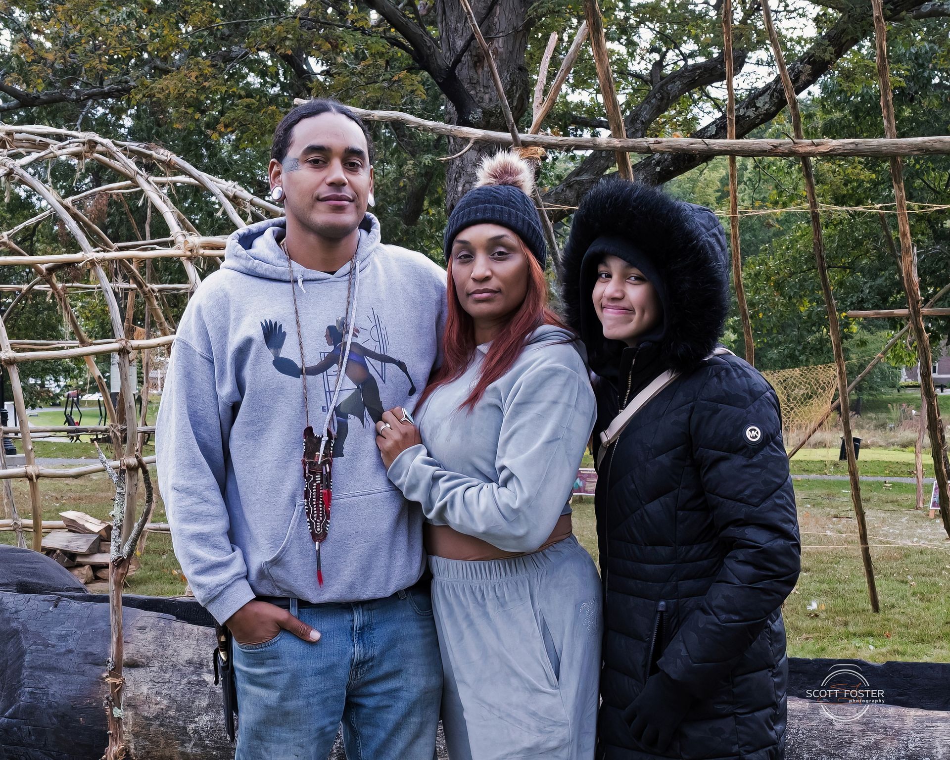 Three people pose outdoors near a wooden structure. Two women and a person with a gray hoodie.