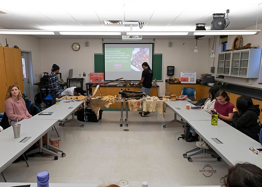 People in a classroom watch a presentation; various items are on tables.