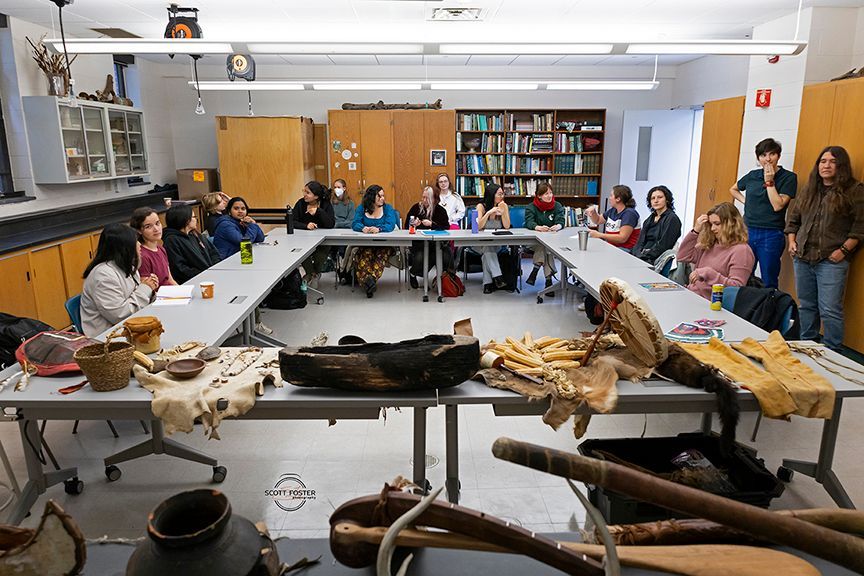 People seated around a table examining artifacts in a classroom, shelves of books in the background.
