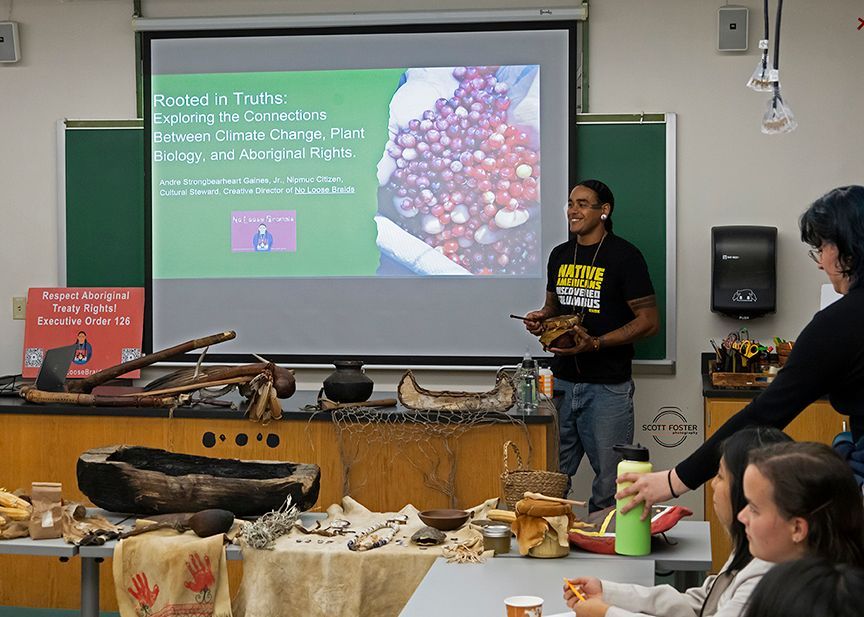 Man giving a presentation about Indigenous topics. Artifacts on a table, projection screen, people watching.
