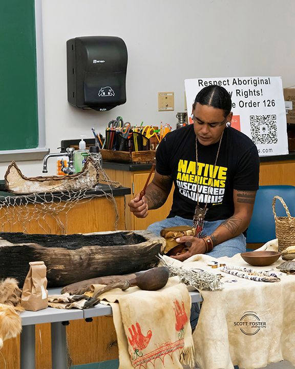 A man with tattoos carves wood at a table with native artifacts. A sign reads, 