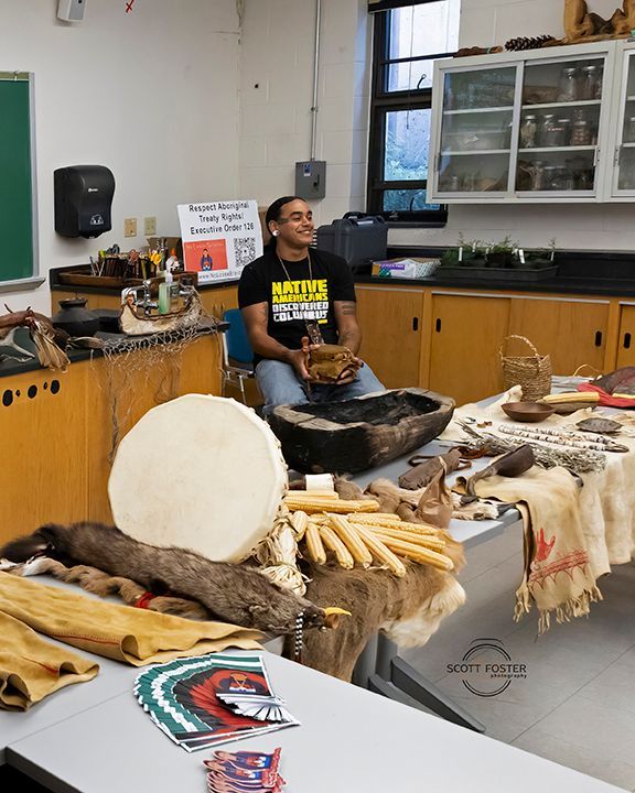 Man sits among cultural artifacts, presenting in a classroom.
