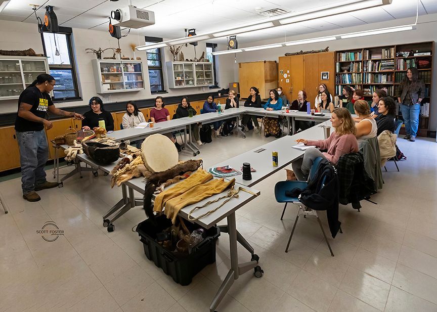 A group of students at tables listens to a presenter. Animal hides and artifacts are displayed. Classroom setting.