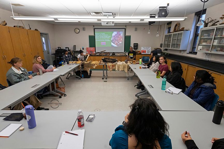 Classroom with students seated around a U-shaped table, listening to a presentation on a screen.