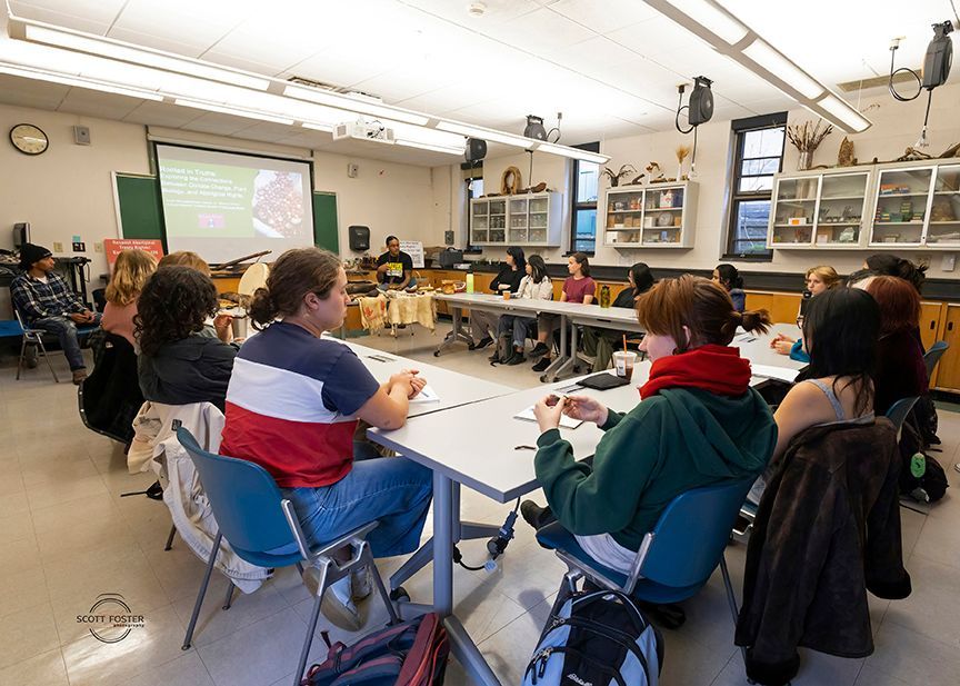 Students in a classroom listen to a presentation. A projector shows an image. Tables are arranged in a square.