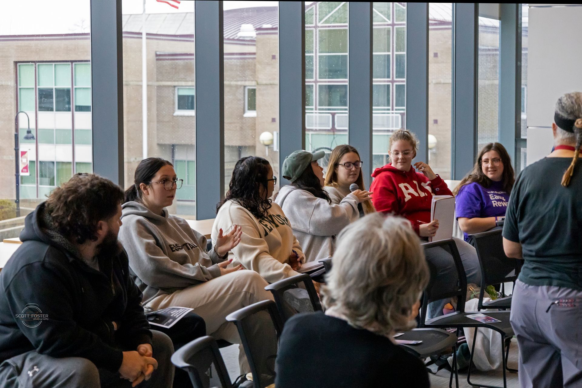 A group of people sitting in chairs in a room with a large window, listening to a speaker.