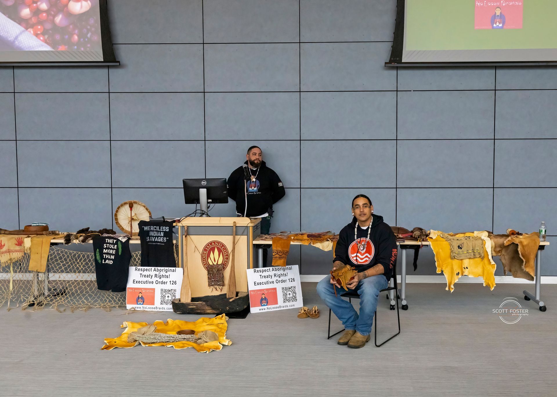 Two men at a display table with furs, crafts, and a computer.
