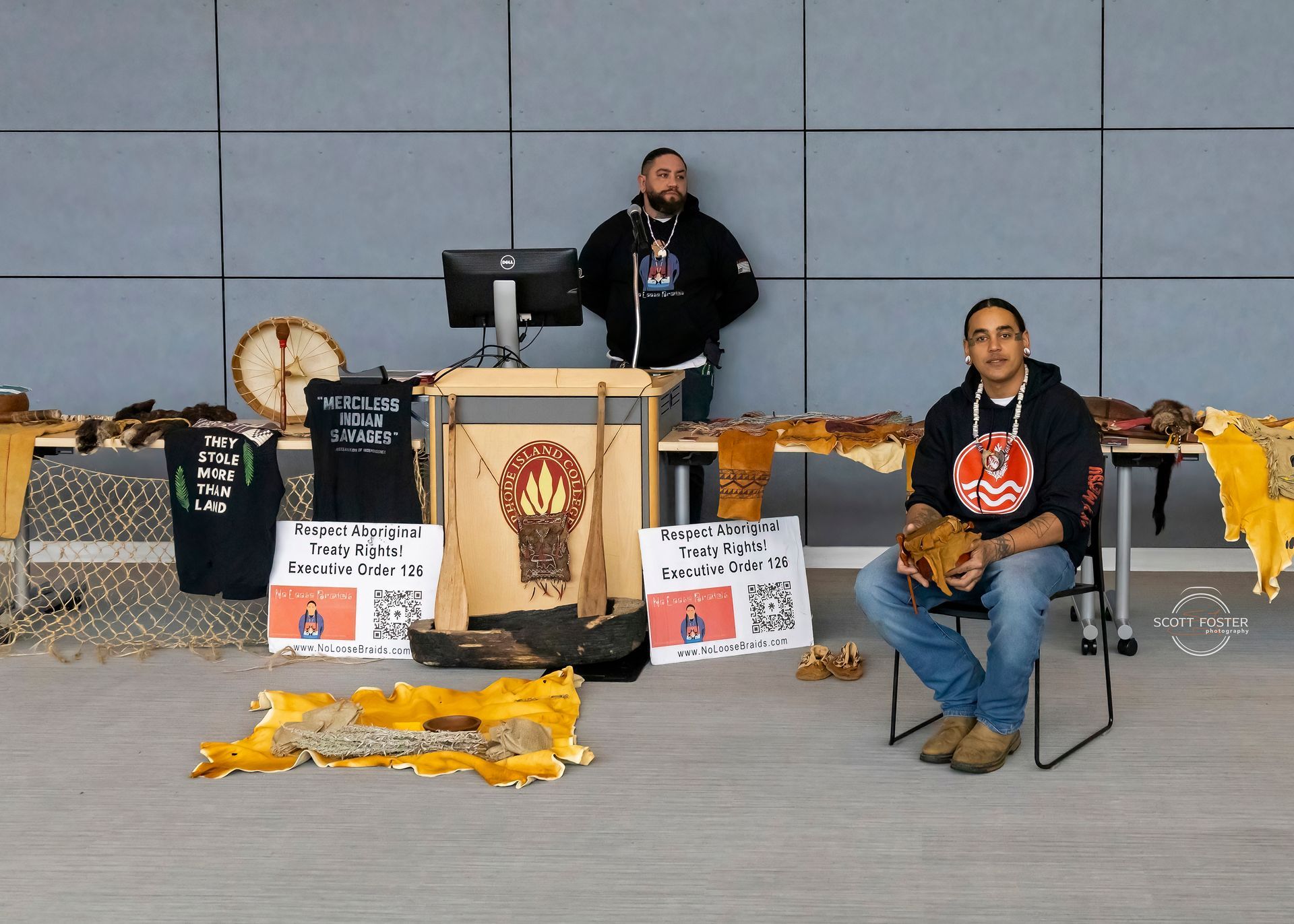 Two men at a display table with Indigenous cultural items, including a drum and furs.