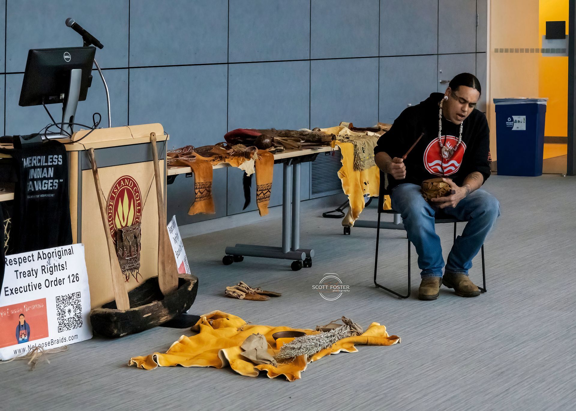 Man seated, crafting leather items. Table with finished pieces, banner, and computer setup. Indoor setting.