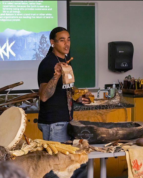 Man presenting with traditional items, standing near table with items and screen.