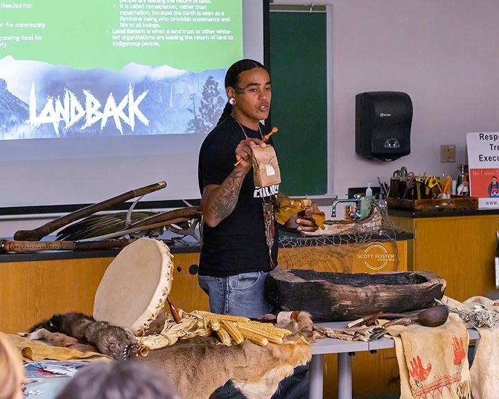 Man speaking, holding items, in front of a table with native artifacts. 