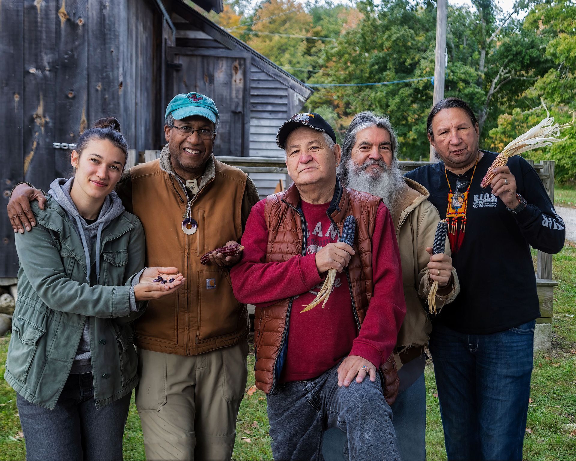 Group of five people smiling outdoors, holding plants, in front of a wooden building.