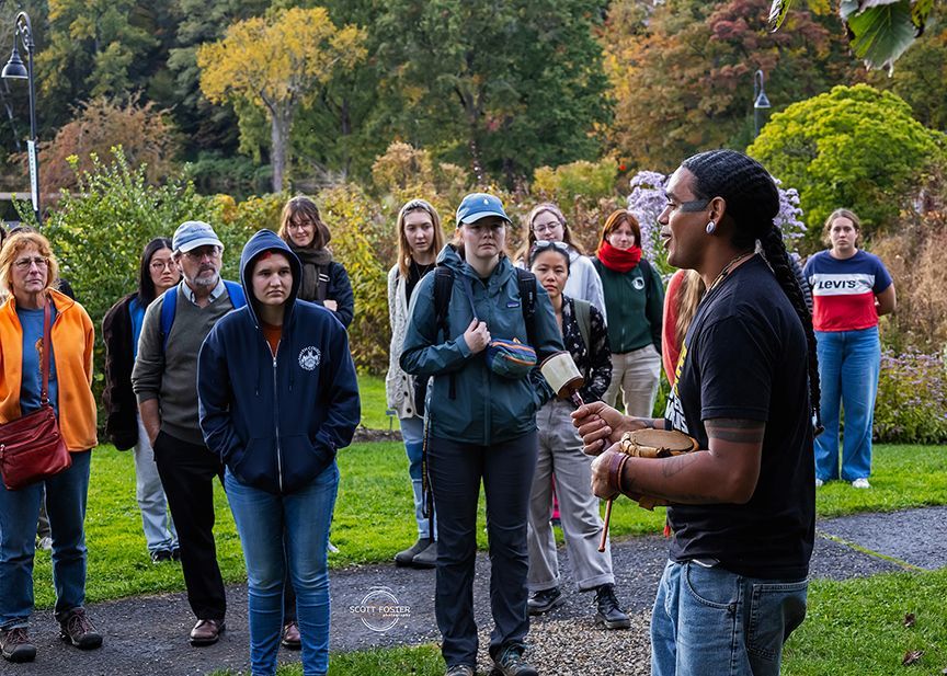 A group of people listens to a man speaking outdoors. Lush greenery and trees are in the background.