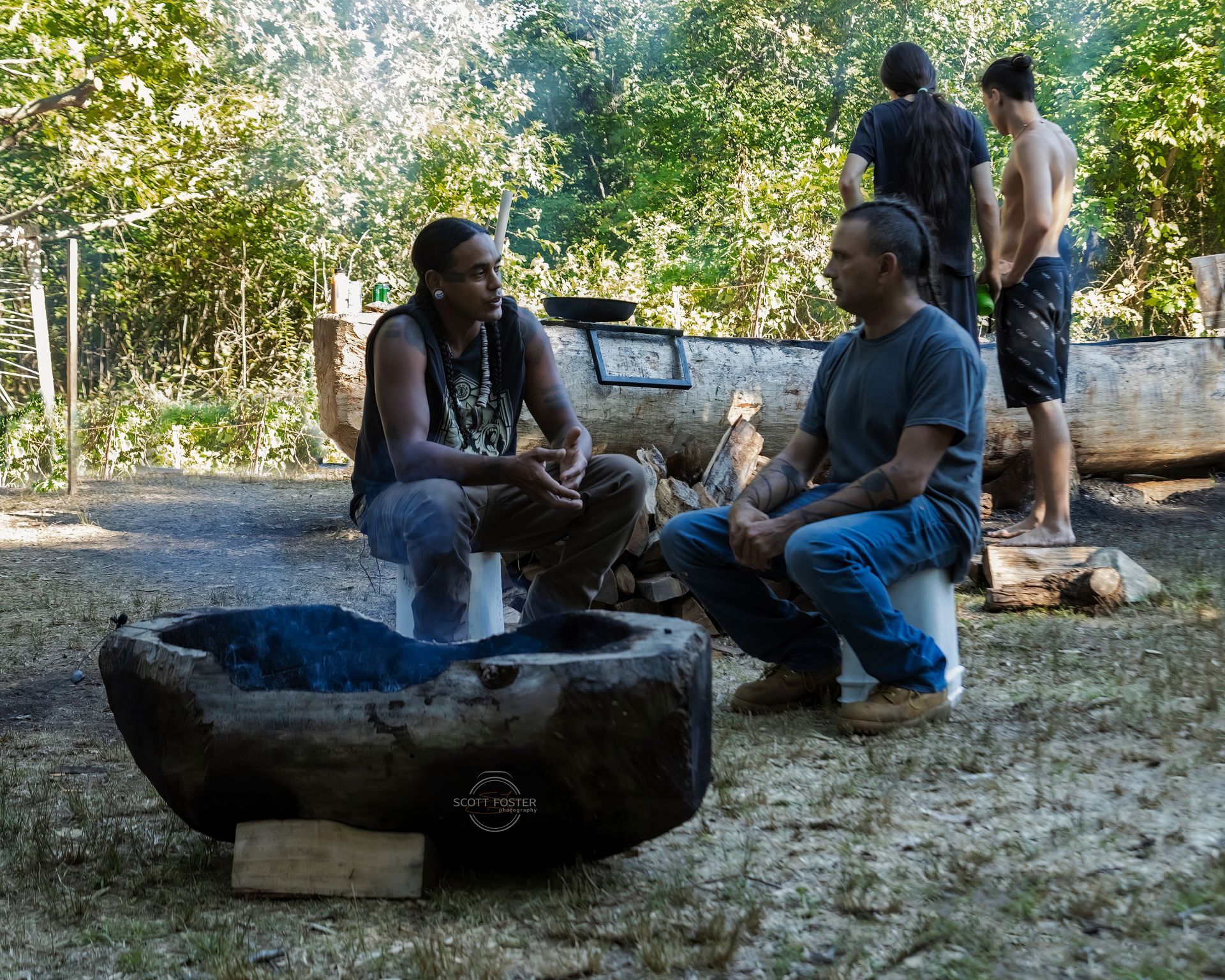 Two men converse near a stone fire pit. Others stand by a large log in a wooded area.