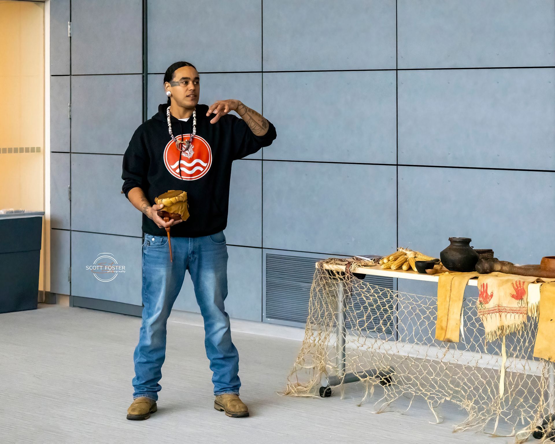 Man in black sweatshirt with red logo, gesturing during presentation, holding object, table with artifacts.