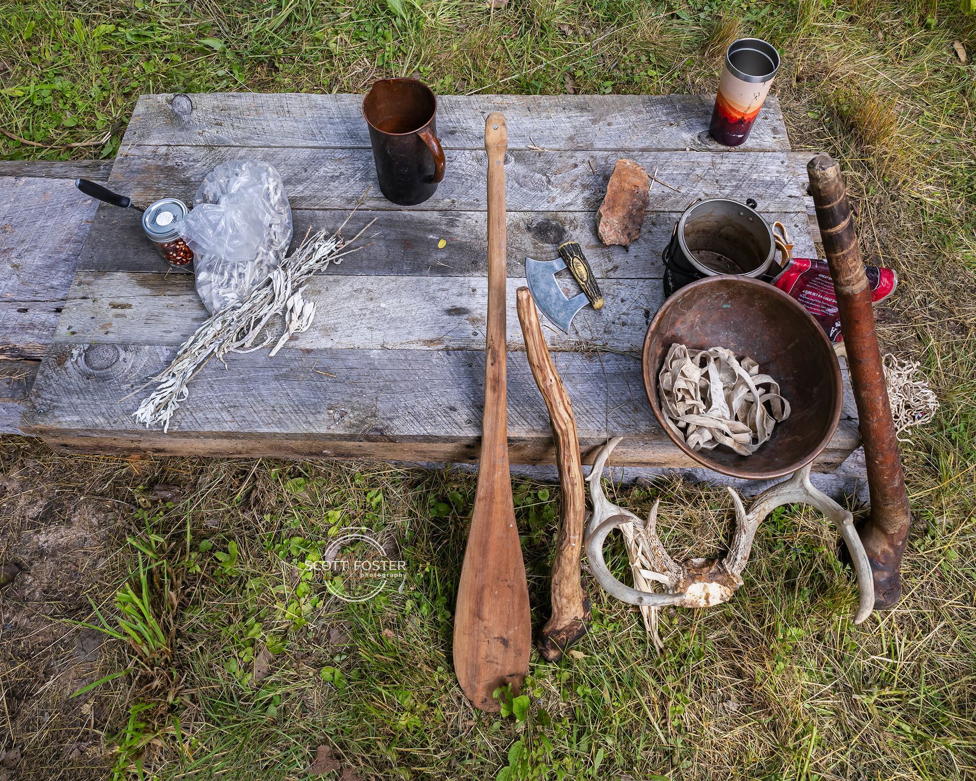 Various rustic objects, including tools and a deer skull, are arranged on a wooden surface outdoors on grass.