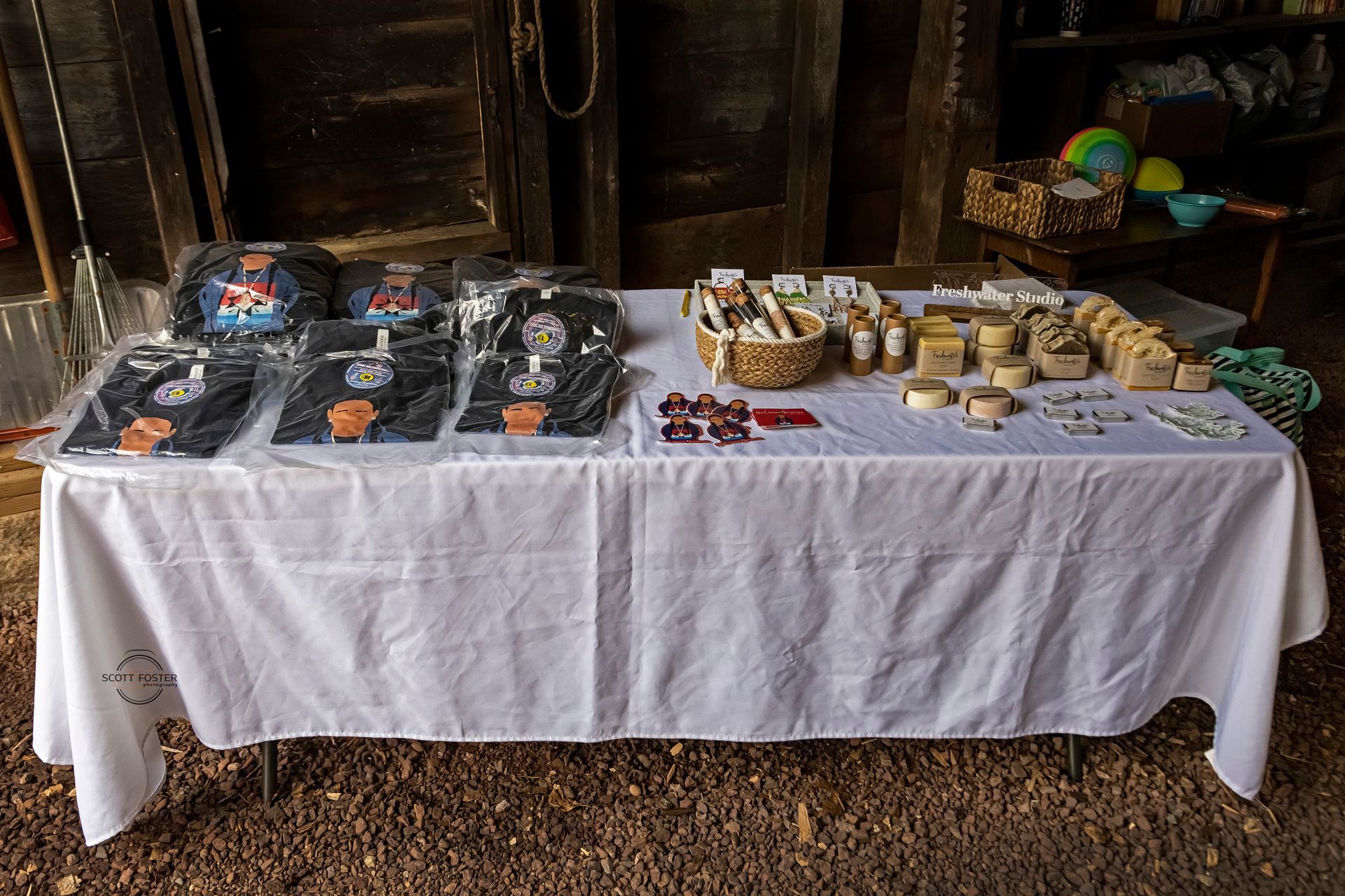 Table of goods for sale on a white tablecloth, indoors. Includes shirts, soaps, and other crafts.