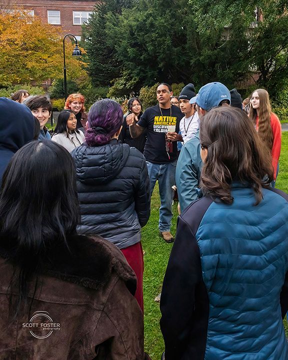 Group of people listening to a person speaking outdoors on a grassy area, buildings in background.