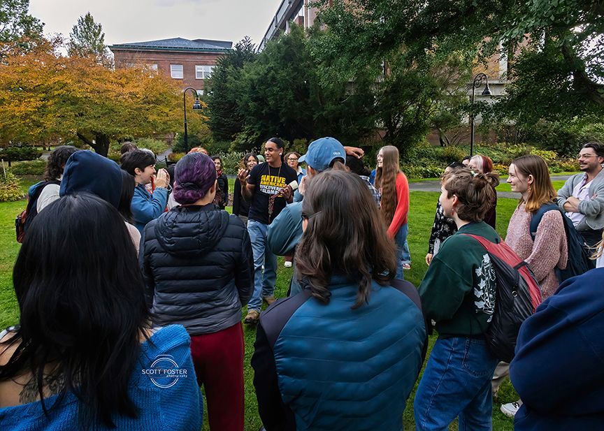 Group of people gathered outdoors, listening to a person speaking. Autumn trees and a building are in the background.