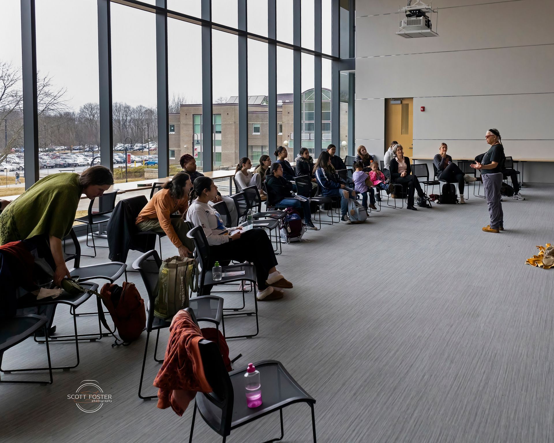 A person speaks to a seated group in a room with large windows, possibly a lecture or presentation.