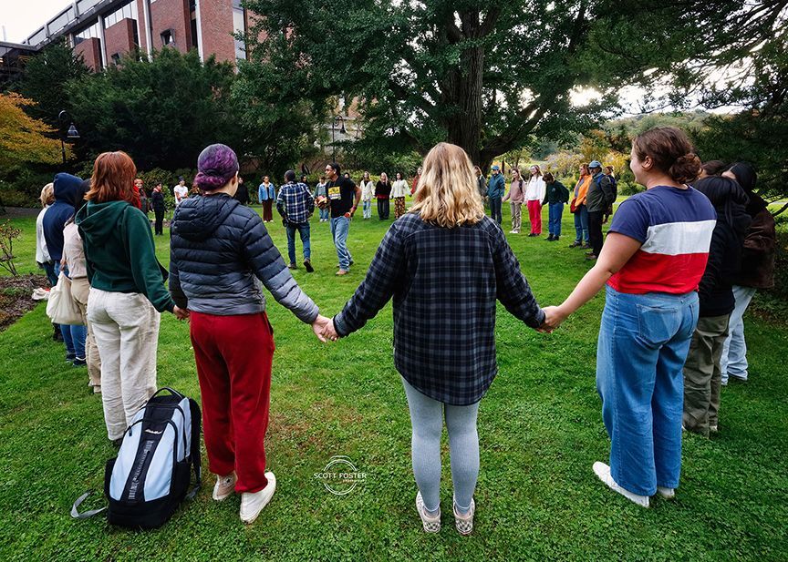 People holding hands in a circle on a grassy area, trees in the background.