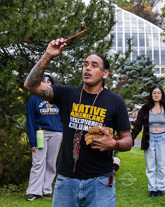 Man with tattoos holds a stick and cloth, leading ceremony. Others watch in a garden with a greenhouse in the background.