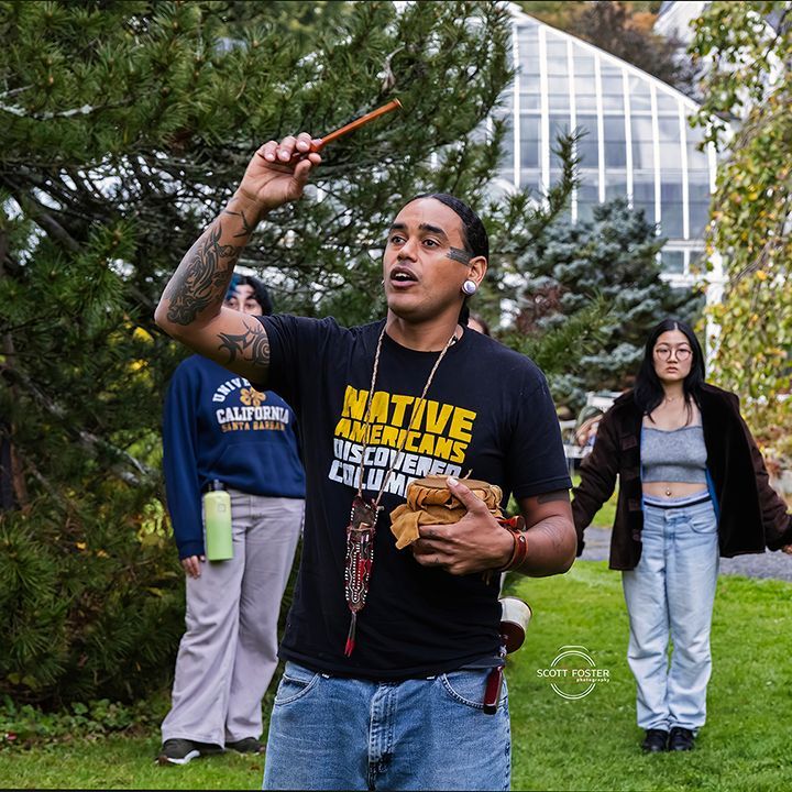 Person holding a stick, gesturing upwards, leading a Native American ceremony outdoors. Other people are in the background.