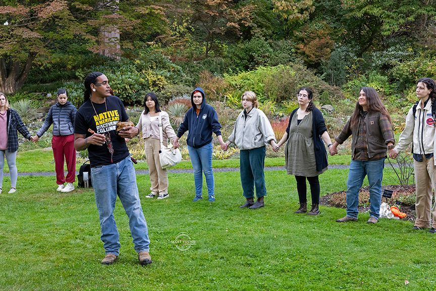 A group of people holding hands in a circle on grass, led by a man speaking. Outdoors, trees in the background.