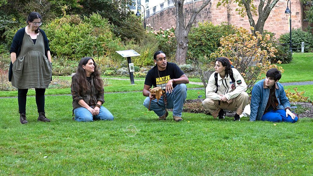 Five people in a grassy area, some squatting. A woman stands, others kneel or crouch. 
