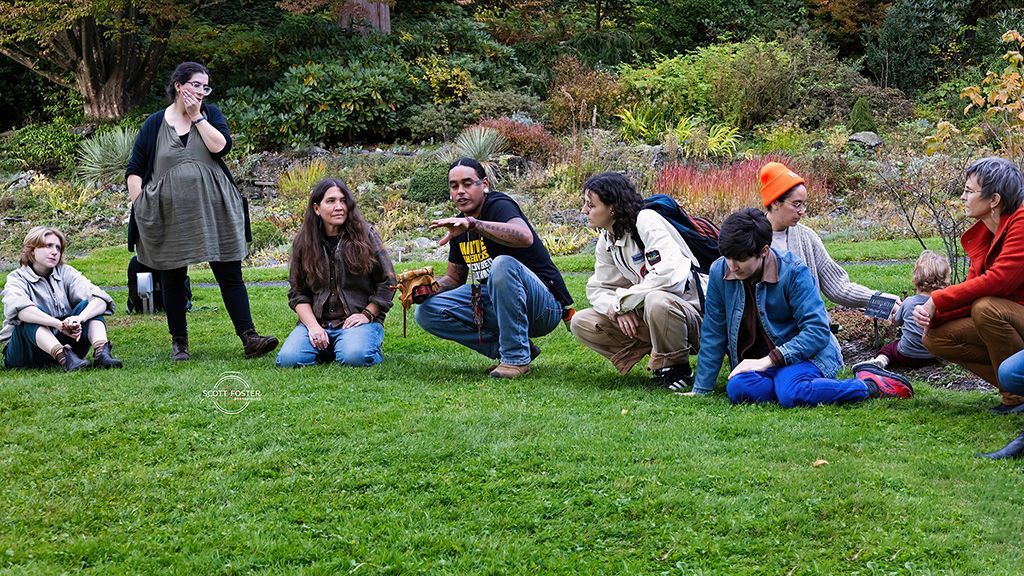 Group of people squatting and standing on grass in a park, one gesturing, others looking on.