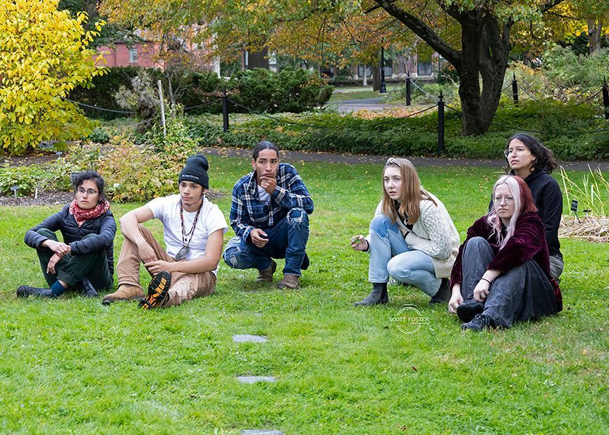 Six people squatting on grass in a park. Trees and foliage in the background. Autumn colors are present.