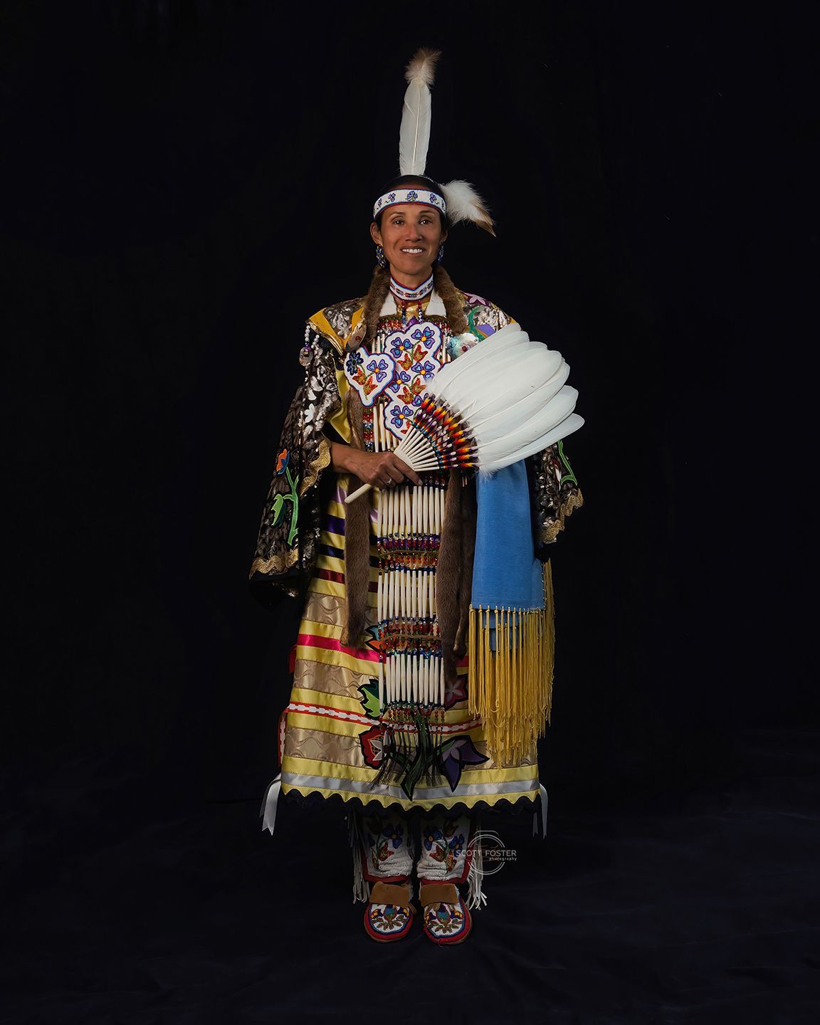 Person in ornate Native American regalia, holding a fan, smiling in front of a black backdrop.