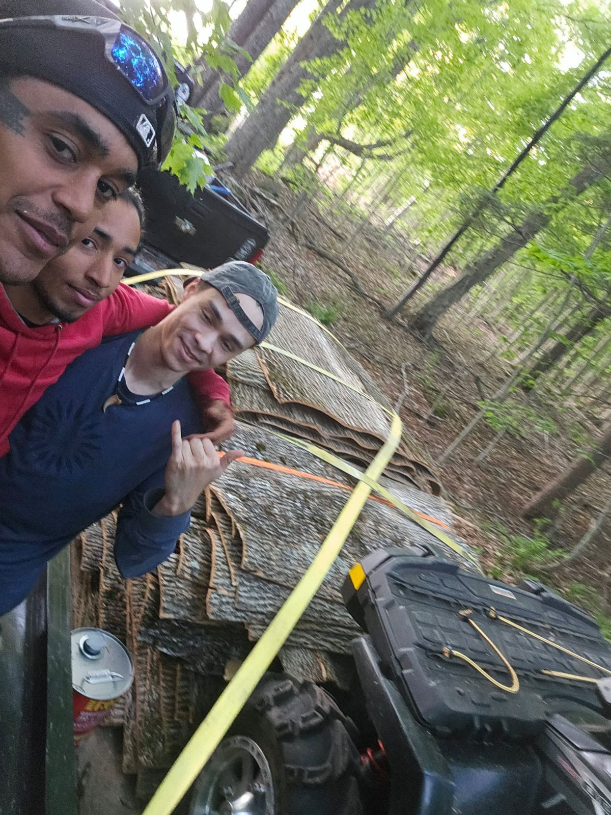 Three men pose near an ATV, large tree, and forest. One man has arm around another.
