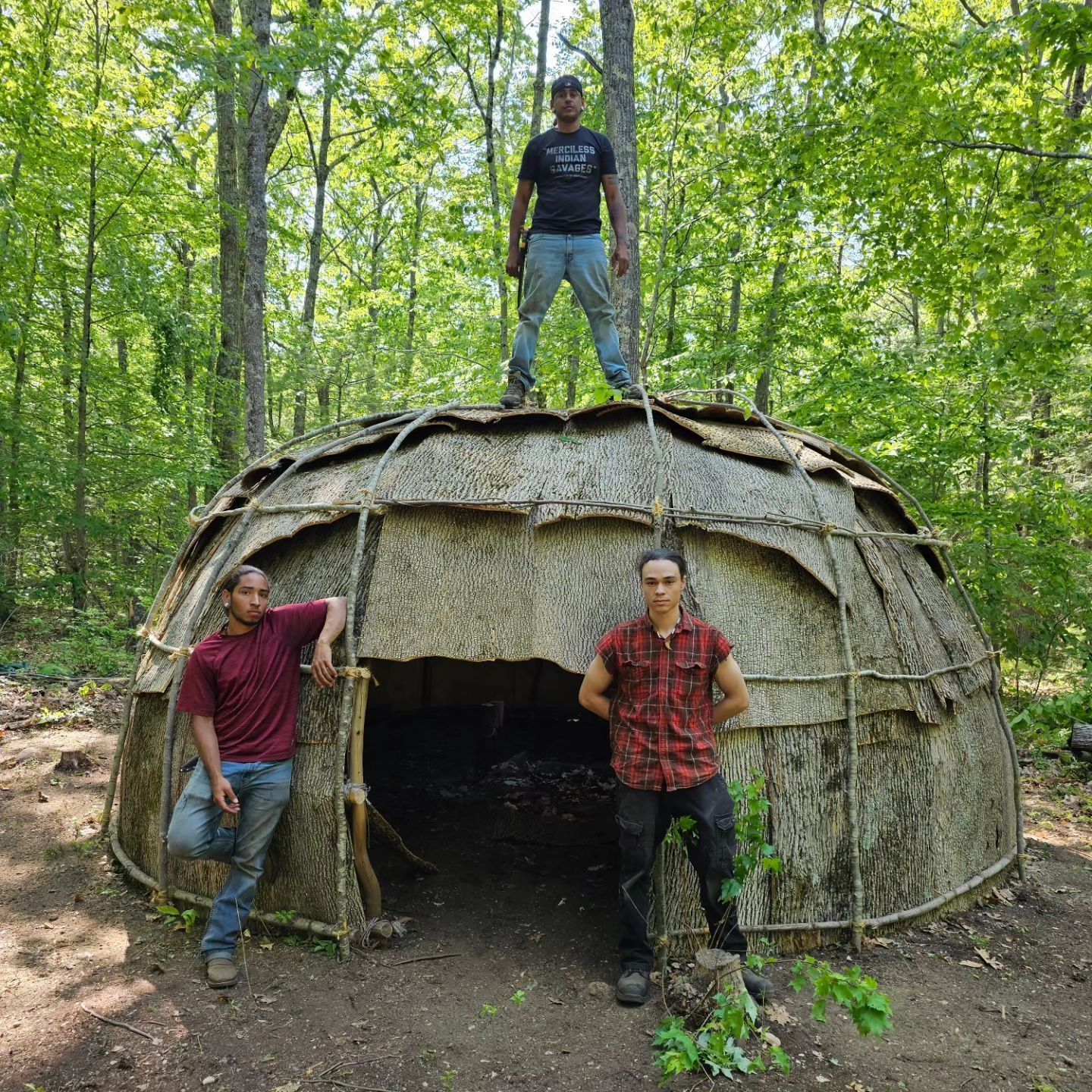 Three people pose near a rustic, domed shelter in a forest. One stands on the roof.