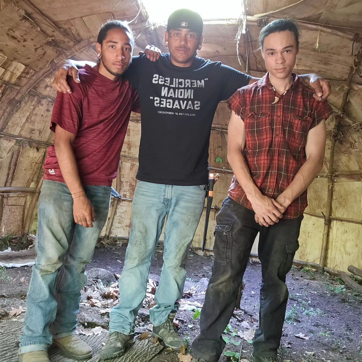Three men inside a wooden structure. Center man with arms around others, wearing a black shirt. Others in jeans and workwear.